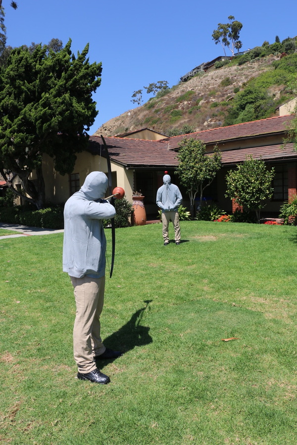 Hooded figure with a bow facing a second figure on the City Hall lawn, Laguna Beach, 2019